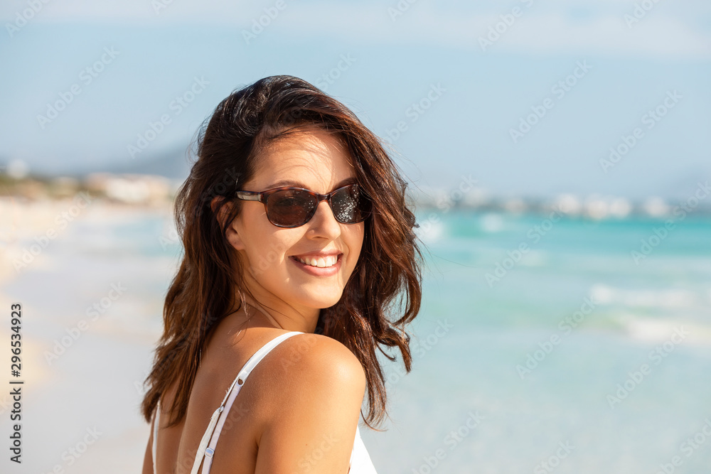 young brunette woman wearing white dress and sunglasses, walking on a beach at the mediterranean sea, or Caribbean 