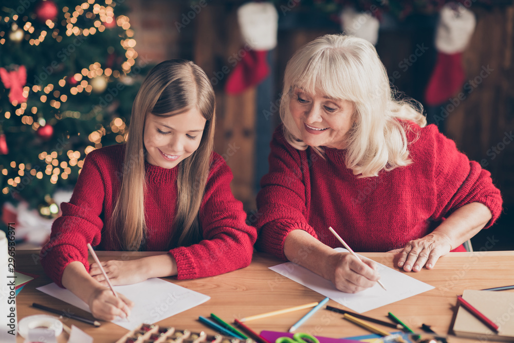Fototapeta premium Portrait of nice attractive lovely inspired cheerful granny pre-teen grandchild writing festal wish letter to Santa Christmastime at decorated industrial brick wood loft style lights interior house
