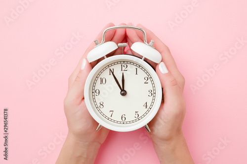 Woman hands holding white alarm clock on light pastel pink background. Time concept. Closeup.