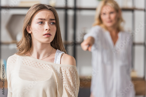 selective focus of offended girl looking away while standing near quarreling mother pointing with finger