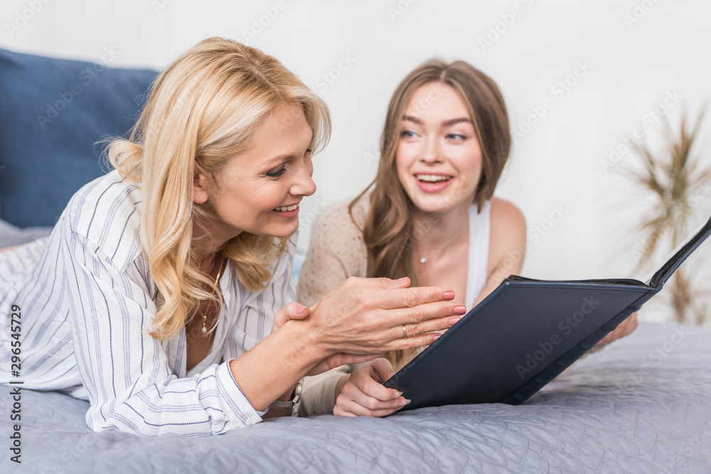 happy woman pointing with hand at photo album while lying on bed near daughter happy woman pointing with hand at photo album while lying on bed near daughter