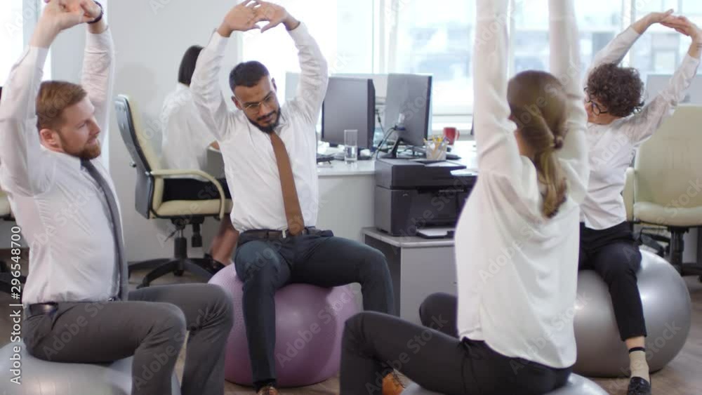 Tilt up of diverse business colleagues sitting on fitballs in office and doing physical exercises together