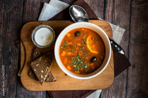 Solyanka soup in a white bowl on the dark rustic wooden table with slices of dark bread and sour cream