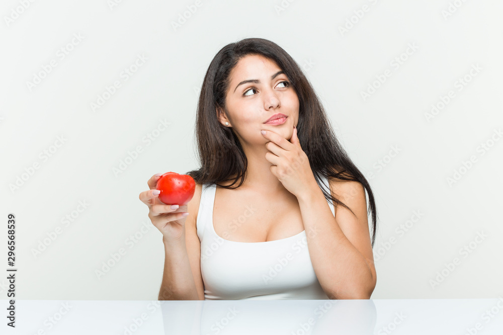 Young hispanic woman holding a tomato looking sideways with doubtful and skeptical expression. Young hispanic woman holding a tomato looking sideways with doubtful and skeptical expression.