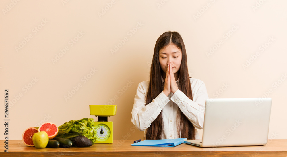 Young nutritionist chinese woman working with her laptop holding hands in pray near mouth, feels confident.