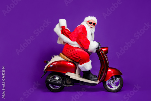Profile photo of fat santa role man rushing newyear party by bike private presents courier delivery wear sun specs and red x-mas costume isolated purple background