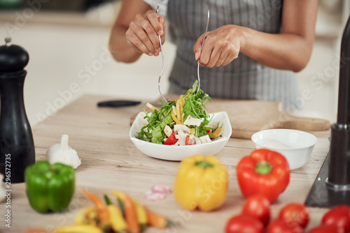 Close up of mixed race woman in apron mixing vegetables in bowl while standing in kitchen at home.