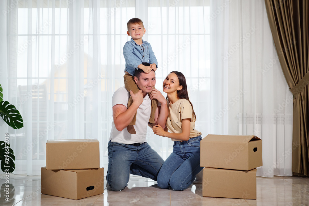 happy smiling family against the background of panoramic windows. Son sits on dad's shoulders near beautiful mother among cardboard boxes. happy smiling family against the background of panoramic windows. Son sits on dad's shoulders near beautiful mother among cardboard boxes.