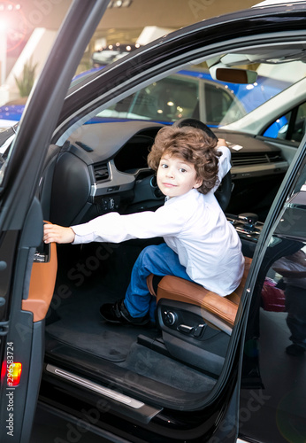Child observing and testing new car in dealership. Little boy sitting in driver's seat, car cabin. Little boy closing the door with his hand.