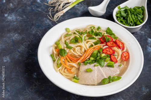 Asian soup with wheat noodles, slices of meat, red chili peppers. Decorated with mint leaves, basil, green onion feathers. There are carrots and broccoli. A healthy, dietary product. Copy space.  