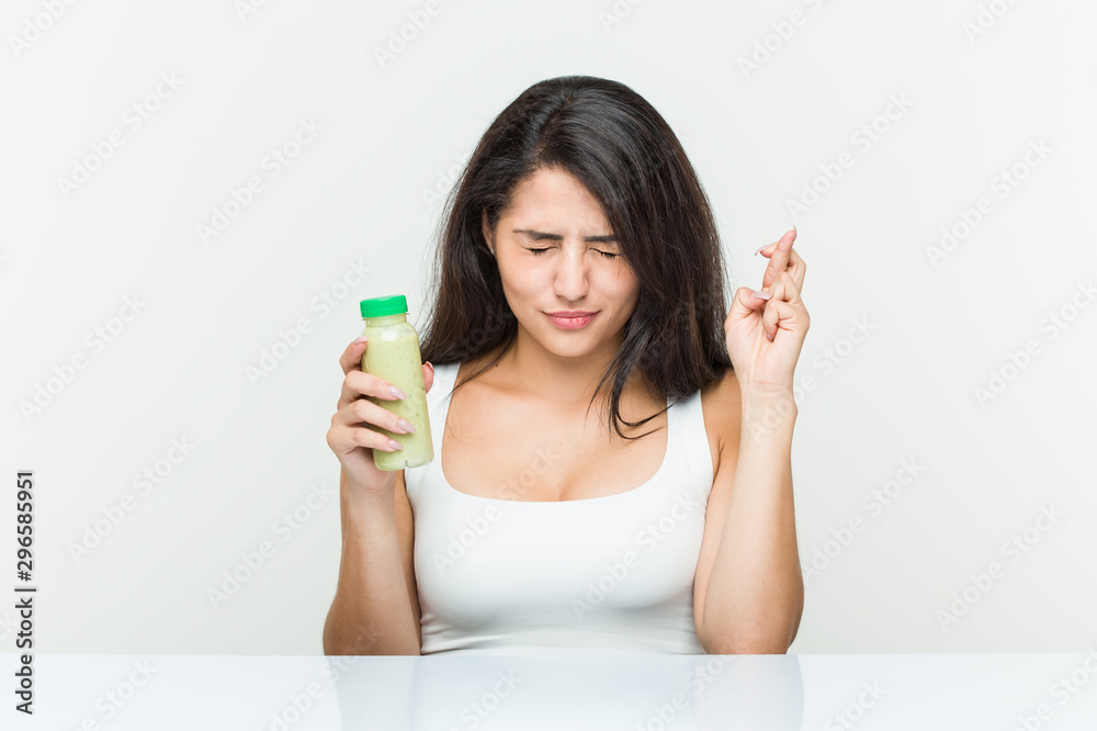 Young hispanic woman holding a vegetable drink crossing fingers for having luck Young hispanic woman holding a vegetable drink crossing fingers for having luck