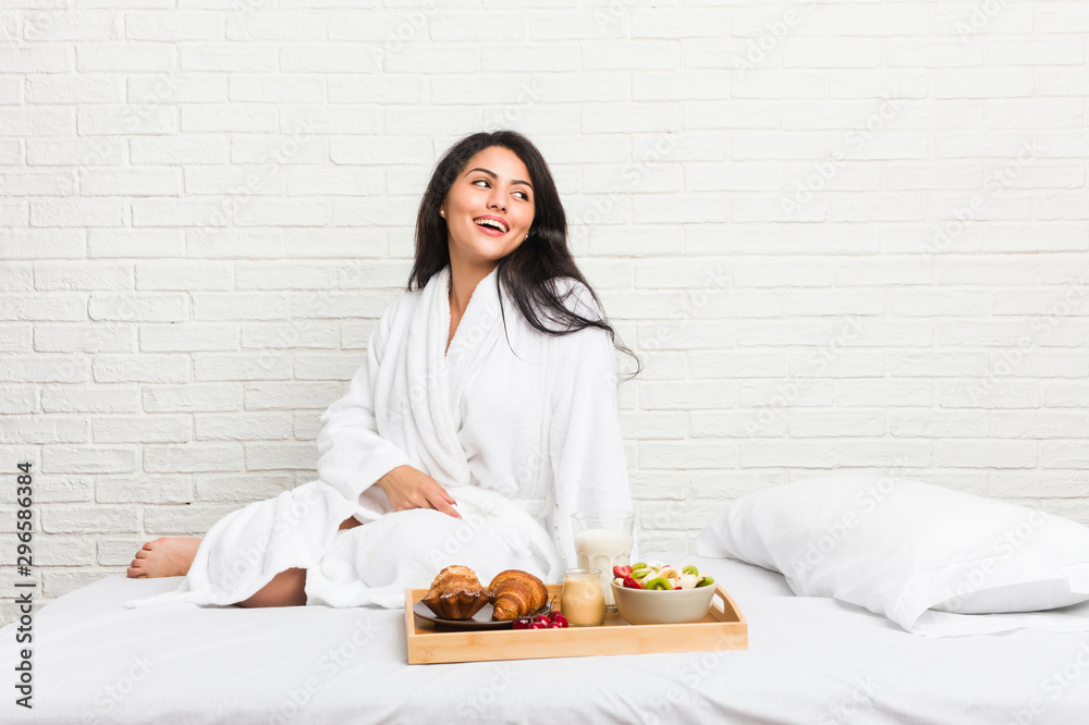 Young curvy woman taking a breakfast on the bed looks aside smiling, cheerful and pleasant.
