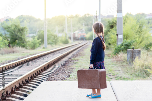 Beautiful charming little girl with pigtails waiting for train at station dressed dark blue dress with flowers and blouse holding big vintage luggage. Young traveler, retro stylization. Cute kid