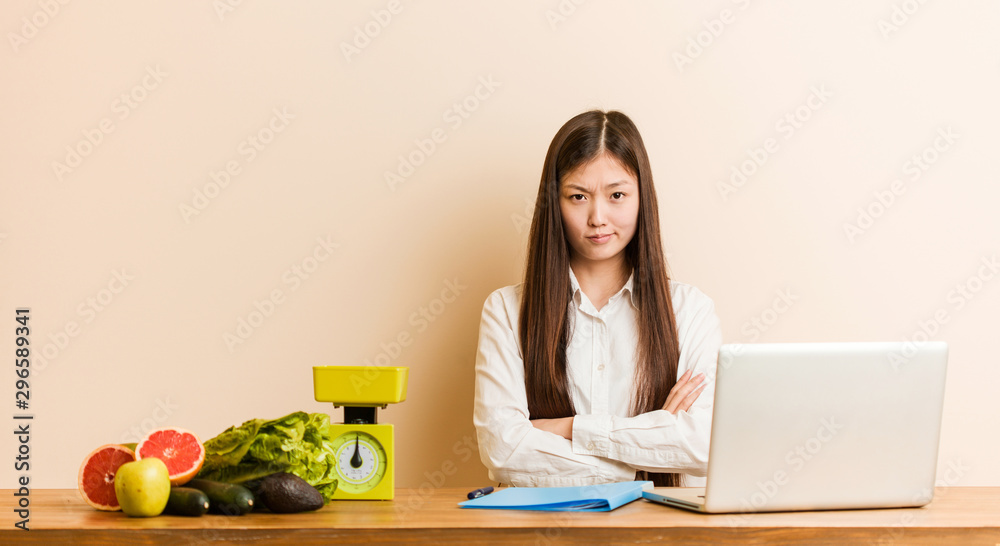 Young nutritionist chinese woman working with her laptop frowning face in displeasure, keeps arms folded. Young nutritionist chinese woman working with her laptop frowning face in displeasure, keeps arms folded.