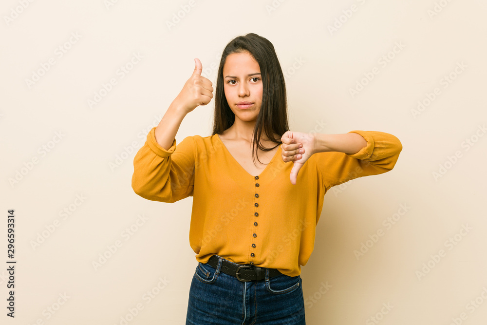 Young hispanic woman against a beige background showing thumbs up and thumbs down, difficult choose concept Young hispanic woman against a beige background showing thumbs up and thumbs down, difficult choose concept