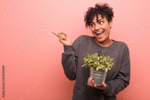 Young african american woman holding a plant smiling cheerfully pointing with forefinger away.