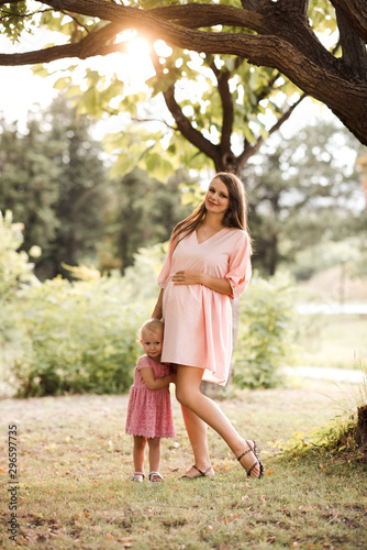 Smiling pregnant woman holding baby girl standing in park over nature background in sun light. Looking at camera. Motherhood. Maternity.