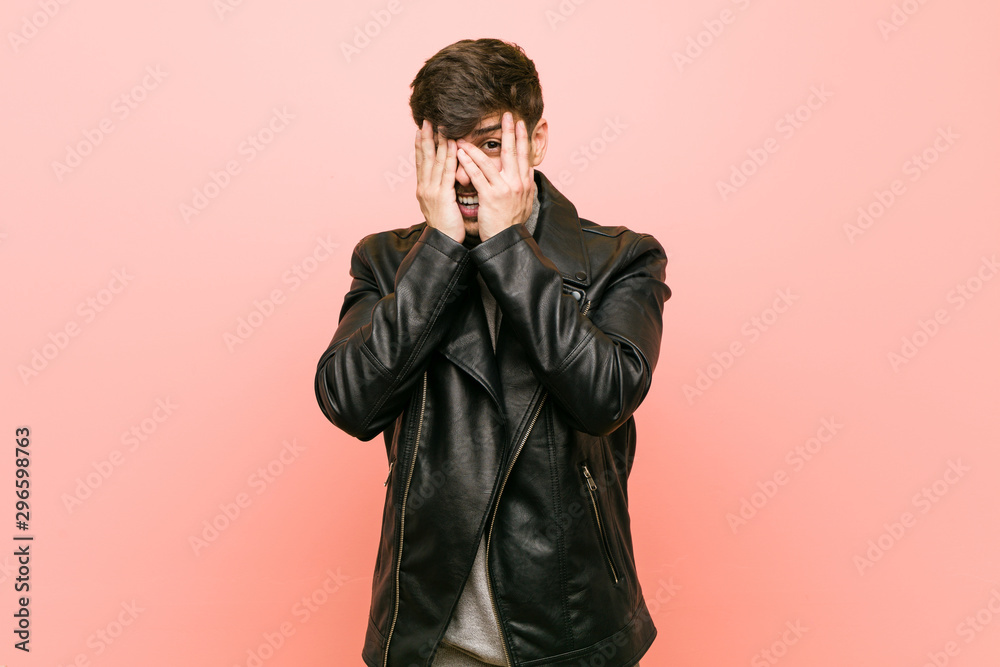 Young hispanic man wearing a leather jacket blink through fingers frightened and nervous.