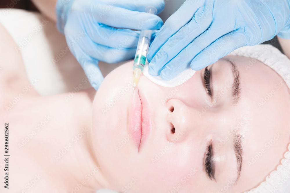Naklejka premium Close-up Attractive young woman gets anti-aging face injections. She lies calmly in a clinic or salon. An experienced young cosmetologist fills female wrinkles with hyaluronic acid from a syringe