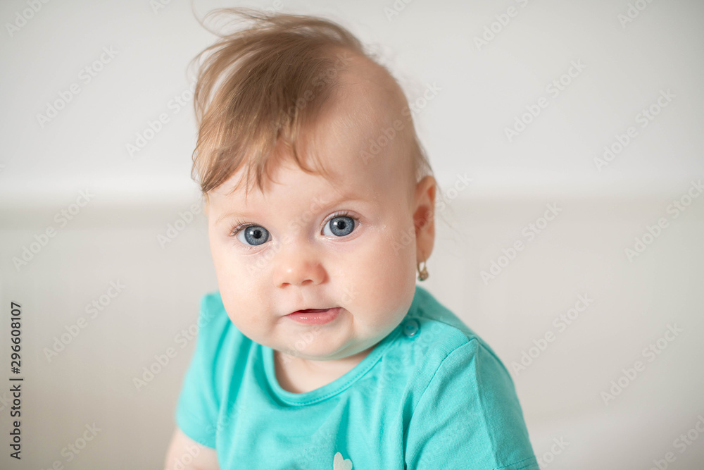 Portrait of adorable Caucasian baby girl with blue eyes, looking at the camera calmly, with curiosity, interactivity or inquisitiveness and sitting in a white baby cot; cute baby expressions concepts