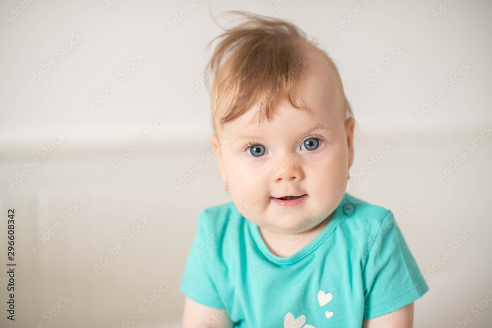 Portrait of adorable Caucasian baby girl with blue eyes, looking at the camera calmly, with curiosity, interactivity or inquisitiveness and sitting in a white baby cot; cute baby expressions concepts