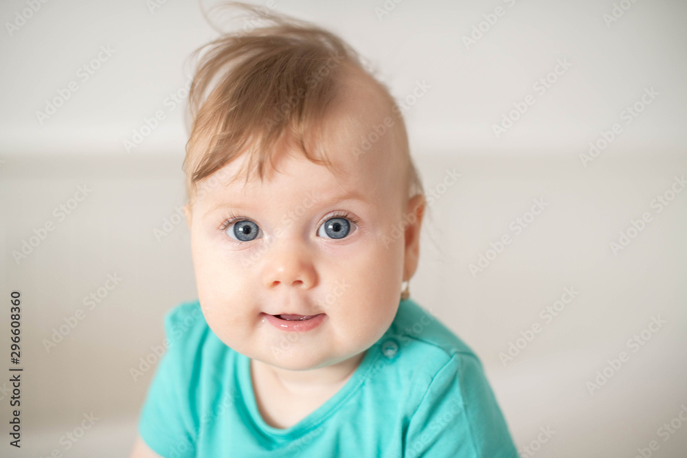 Portrait of adorable Caucasian baby girl with blue eyes, looking at the camera calmly, with curiosity, interactivity or inquisitiveness and sitting in a white baby cot; cute baby expressions concepts