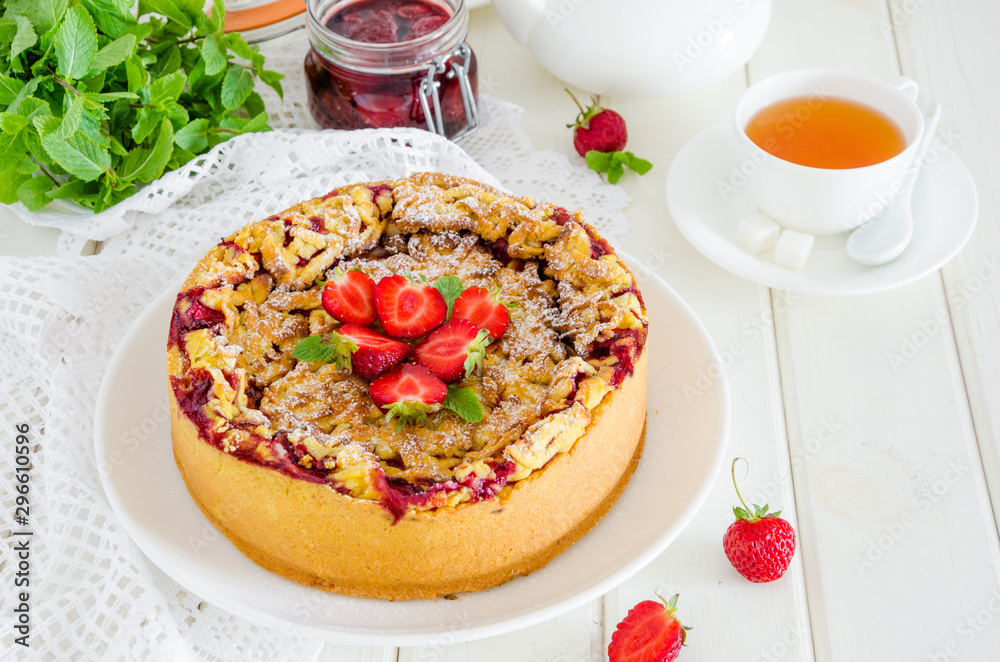 Baked creamy cheesecake with strawberries and crispy shtrezel on a plate on a white wooden background with a cup of tea.