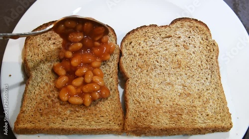Video of spoon filled with baked beans and adding beans on toast. Placing baked beans onto two slices of brown whole meal toast. Toast topped with British baked beans video. Simple breakfast meal.