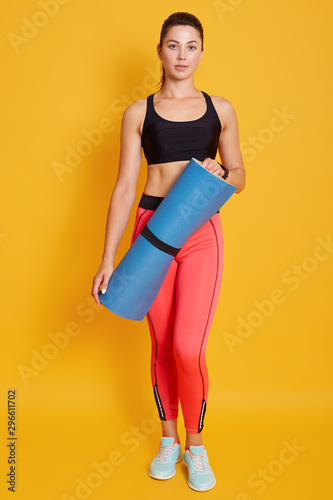 Toned young beautiful dak haired woman athlete wearing sportswear, standing and holding blue fitness mat, looking directly at camera, fit female after yoga session or training. Sport concept.