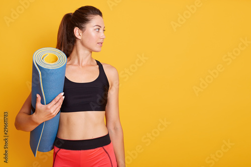 Close up portrait of young beautiful Caucasian woman standing and holding blue yoga mat in hand, ready for exercising in gym, wears stylish top and leggins, looking aside. Copy space for advertisment.