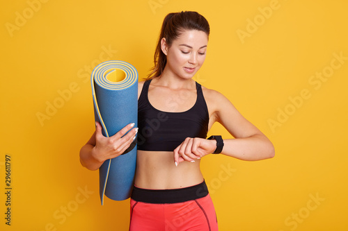 Studio shot of female afer workout session checks results on smartwatch in fitness app, woman looks at smart watch, counts up spent calories after training in gym. Sport and healthy lifestyle concept. photo
