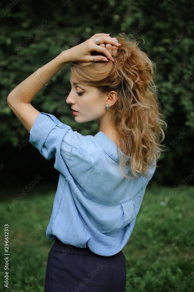 Back view portrait of sexy blonde girl with curly hair wearing blue casual jeans blouse posing in the park on green trees background. Hipster style concept. Back view portrait of sexy blonde girl with curly hair wearing blue casual jeans blouse posing in the park on green trees background. Hipster style concept.