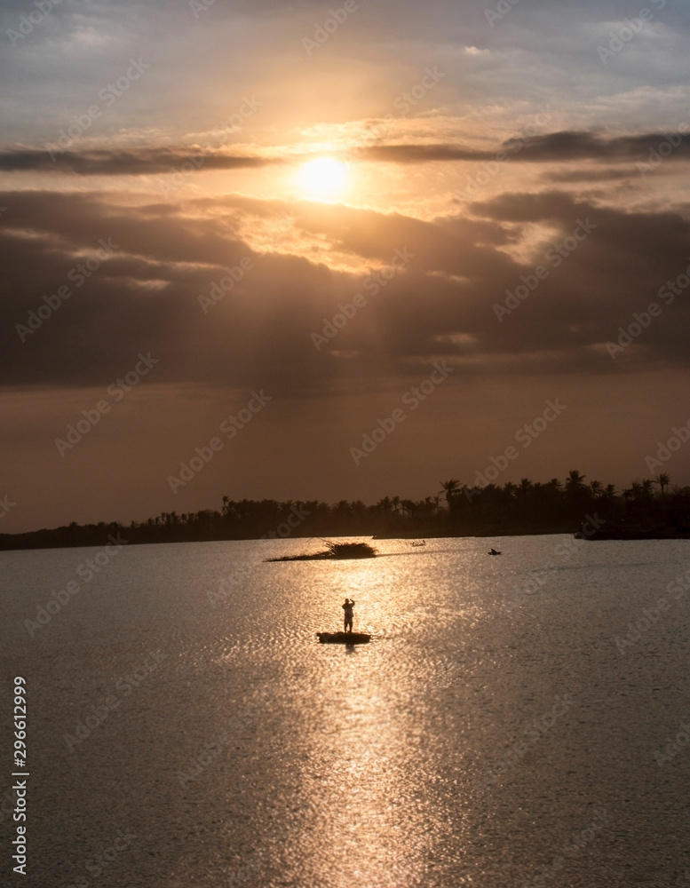 A fisherman in the middle of the sea with a ray of light emerges from the thick clouds above him at sunset