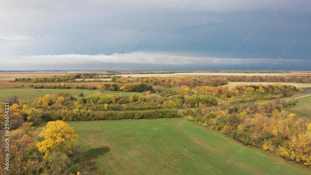 Drone moving slowly forwards to show a beautiful scene with a bright autumn colored forest, farm, and river and landscape in early autumn colors. A road cuts through the scene and storm clouds are on Drone moving slowly forwards to show a beautiful scene with a bright autumn colored forest, farm, and river and landscape in early autumn colors. A road cuts through the scene and storm clouds are on