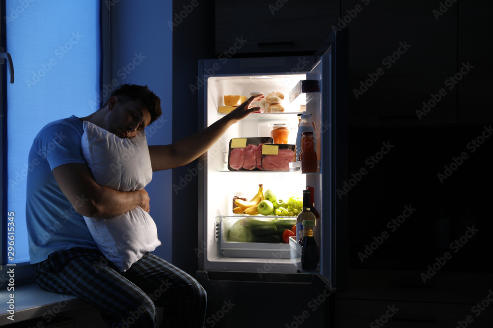 Sleepy man with pillow near refrigerator in kitchen at night