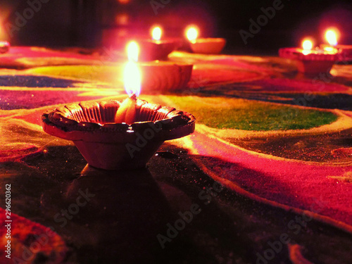 low angle shot of diwali oil clay lamps with intricate design and glowing hazy flame placed on multicoloured green blue red rangoli