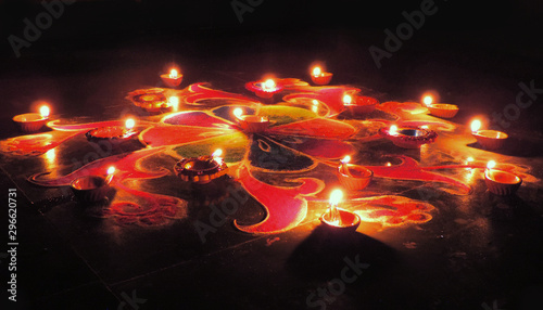 side view of round red blue green Diwali Rangoli pattern with dominant red orange drawn on a dark black background illuminated by oil clay deep lamps