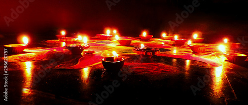 low angle view of round red blue green Diwali Rangoli pattern with dominant red orange drawn on dark black background illuminated by oil clay deep lamps