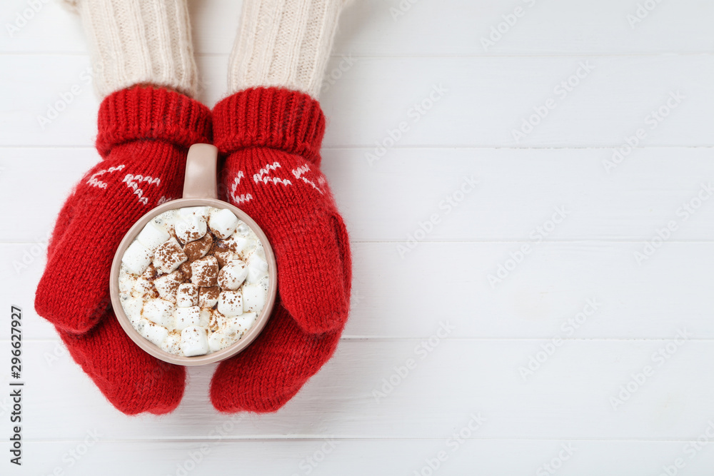 Hands in knitted mittens holding cup of hot drink on white wooden table - obrazy, fototapety, plakaty 