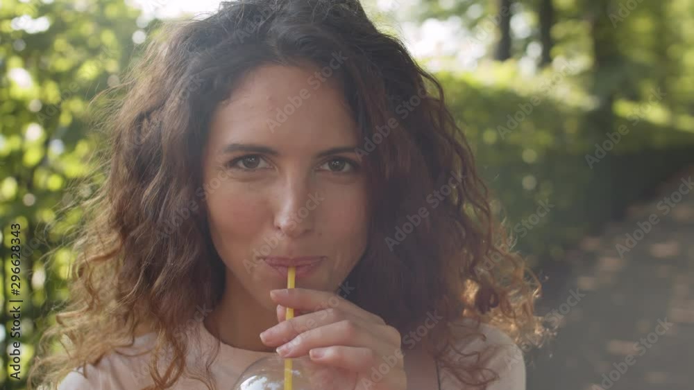 Tracking portrait shot of beautiful young woman with curly hair posing for camera in park on sunny summer day. She is smiling and drinking juice or smoothie Tracking portrait shot of beautiful young woman with curly hair posing for camera in park on sunny summer day. She is smiling and drinking juice or smoothie