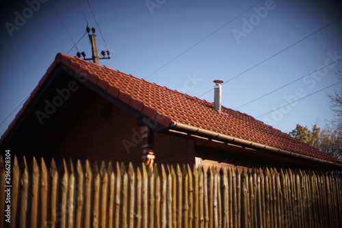 Wooden house with red tiled roof and blue sky. Fence made of sharp wooden stakes. Blurred.