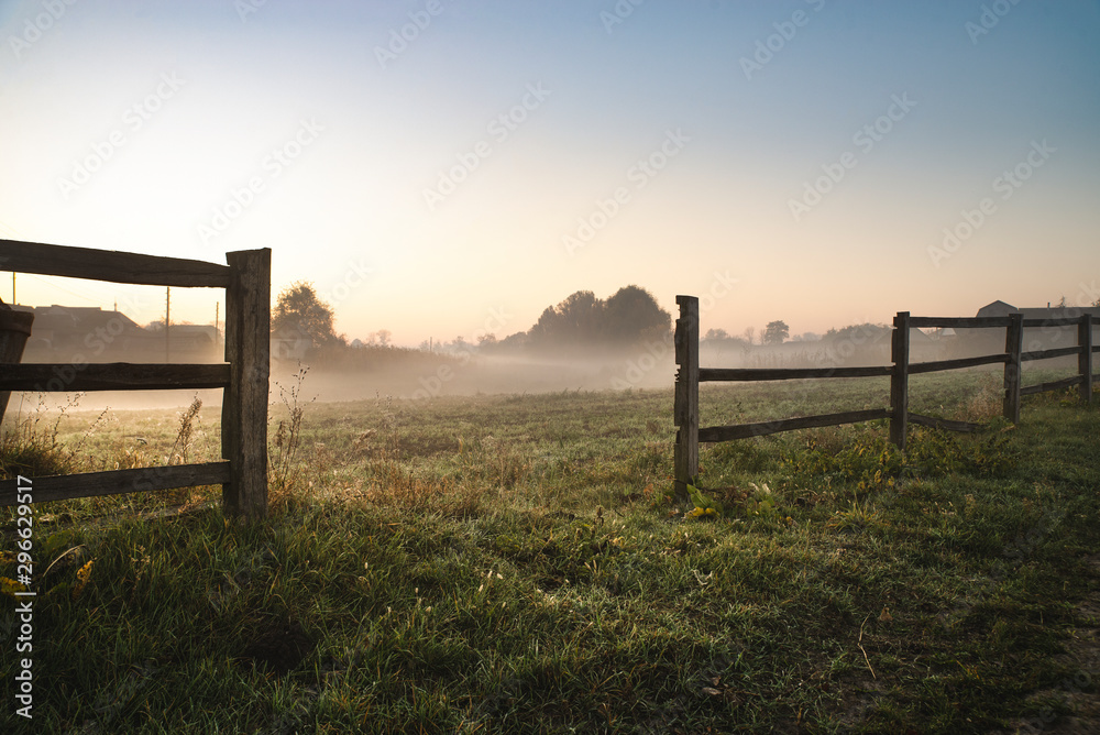 Morning landscape at sunrise. A field with green grass is covered with fog. In the foreground a wooden fence