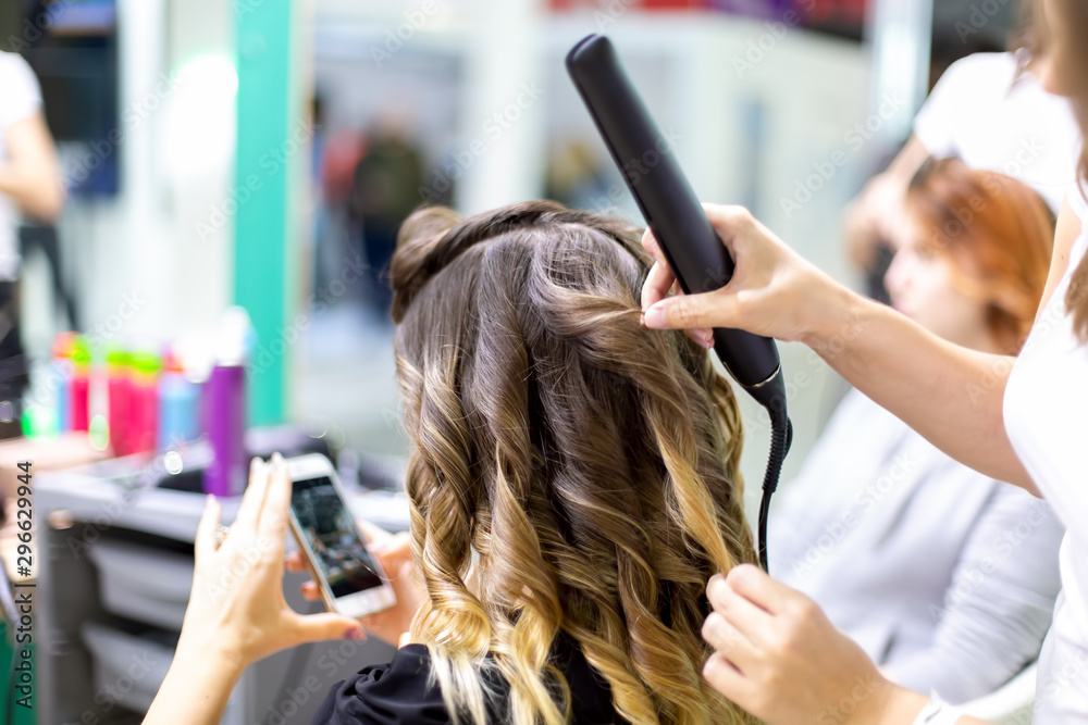 close up stylist makes a girl client styling hair waving with curling irons in a hairdresser. customer girl looks into smartphone in blur. close up, soft focus close up stylist makes a girl client styling hair waving with curling irons in a hairdresser. customer girl looks into smartphone in blur. close up, soft focus