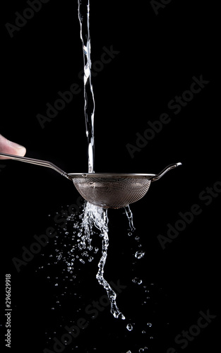 A stream of water pours through a small steel sieve in the hands of a man on a black background. Splashes of water fly out through a sieve.