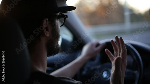 Close-up of young bearded driver, driving with hands on car wheel, wearing glasses and black cap. Talking with passenger.