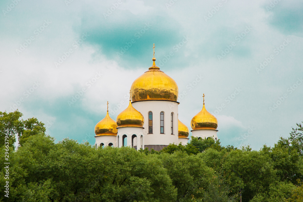golden domes of a church with crosses on a background of blue clear skies. Domes of the church behind green trees on a clear day