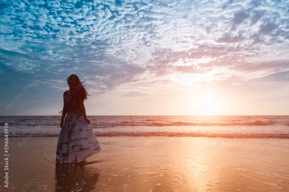 Asian girl wear Indigo dyed cotton long skirt,smiling and flower tucked behind her ear on the beach,blue sky and sea,orange light flare. Asian girl wear Indigo dyed cotton long skirt,smiling and flower tucked behind her ear on the beach,blue sky and sea,orange light flare.