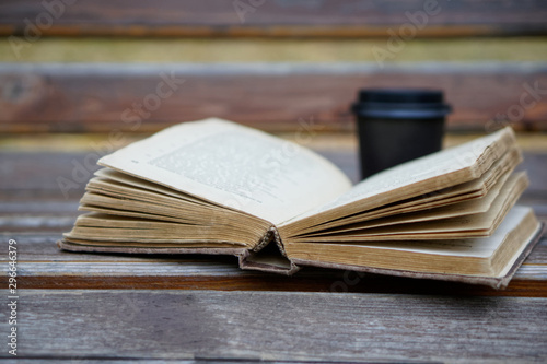 An old shabby thick book lies on a wooden bench next to a cup of coffee
