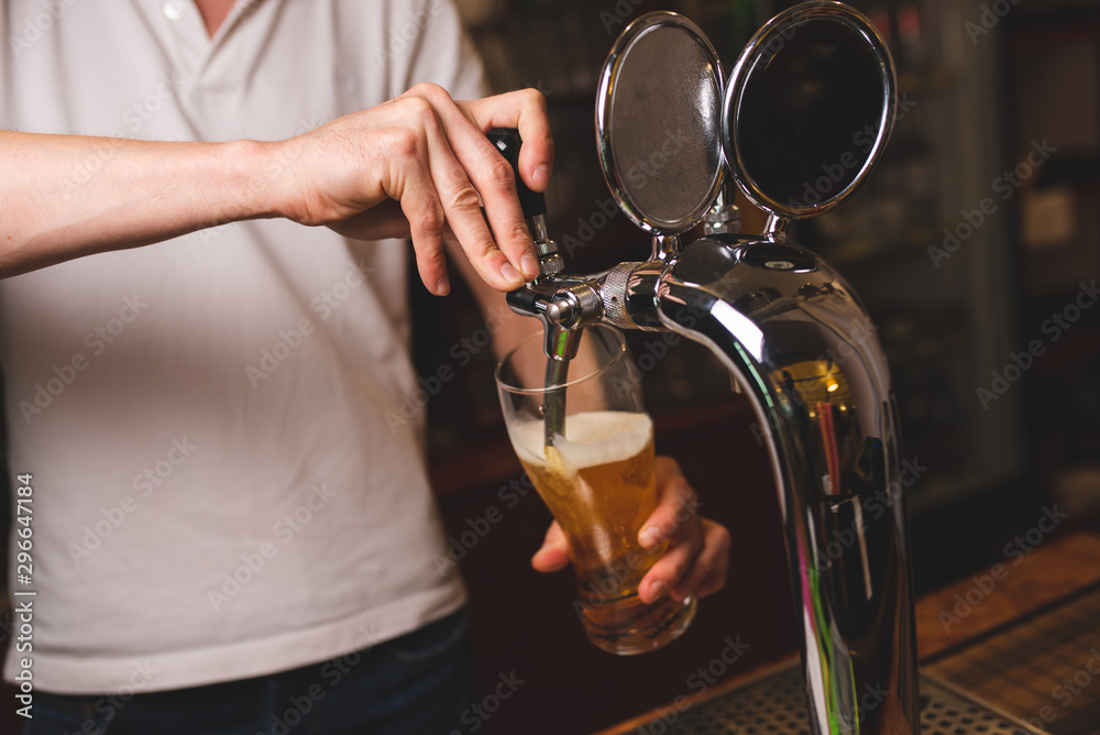 A male barman in a white T-shirt pours a glass of beer from a tap