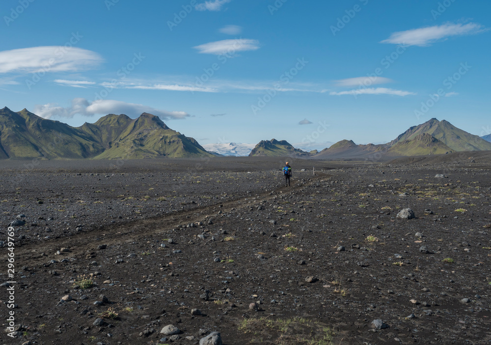 Icelandic lava desert landscape with man hiker on Laugavegur hiking trail with view on Tindfjallajokull glacier mountains and green hills. Fjallabak Nature Reserve, Iceland. Summer blue sky Icelandic lava desert landscape with man hiker on Laugavegur hiking trail with view on Tindfjallajokull glacier mountains and green hills. Fjallabak Nature Reserve, Iceland. Summer blue sky